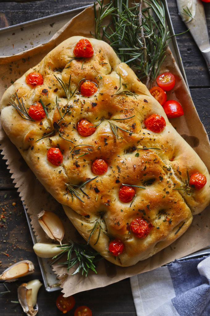Focaccia bread topped with cherry tomatoes, rosemary and olive oil on cloth, with garlic beside